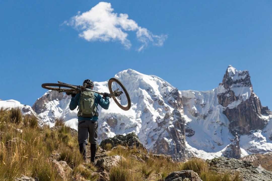 Ciclista cargando la bicicleta al hombro mientras camina por un sendero rocoso de altura, con picos nevados impresionantes bajo cielo azul.