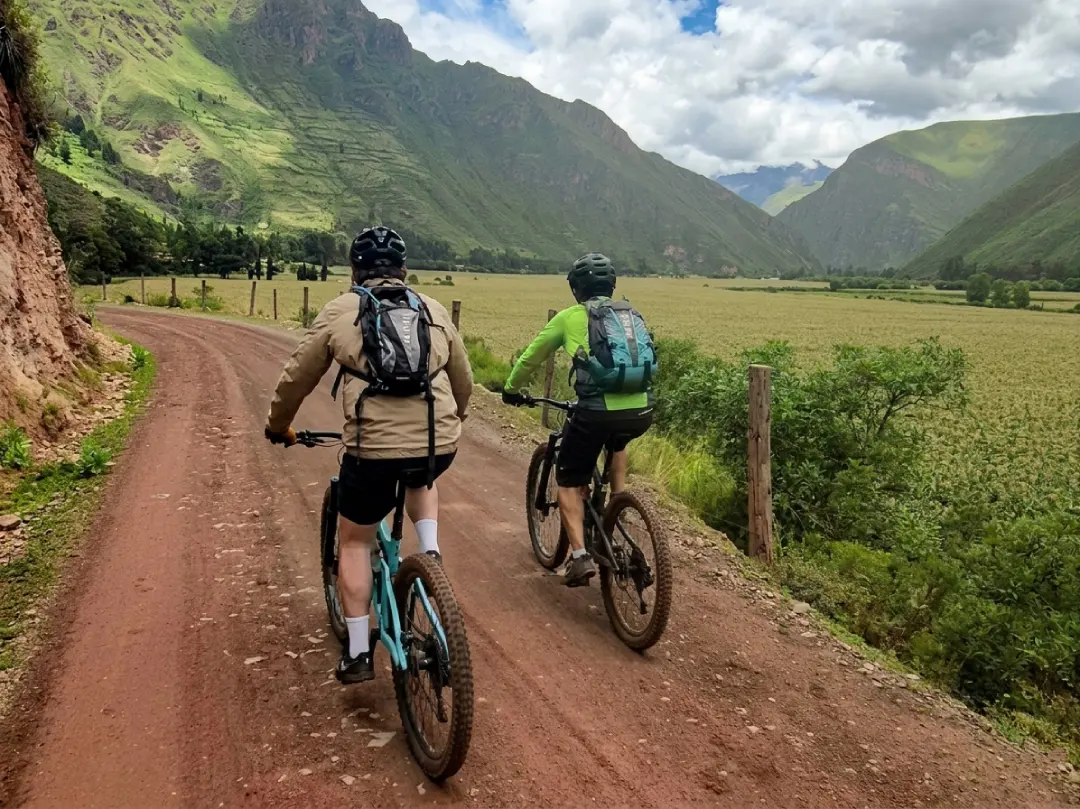 Two cyclists with helmets and backpacks riding on a red dirt road through the Sacred Valley, with green Andean mountains and clouds ahead.