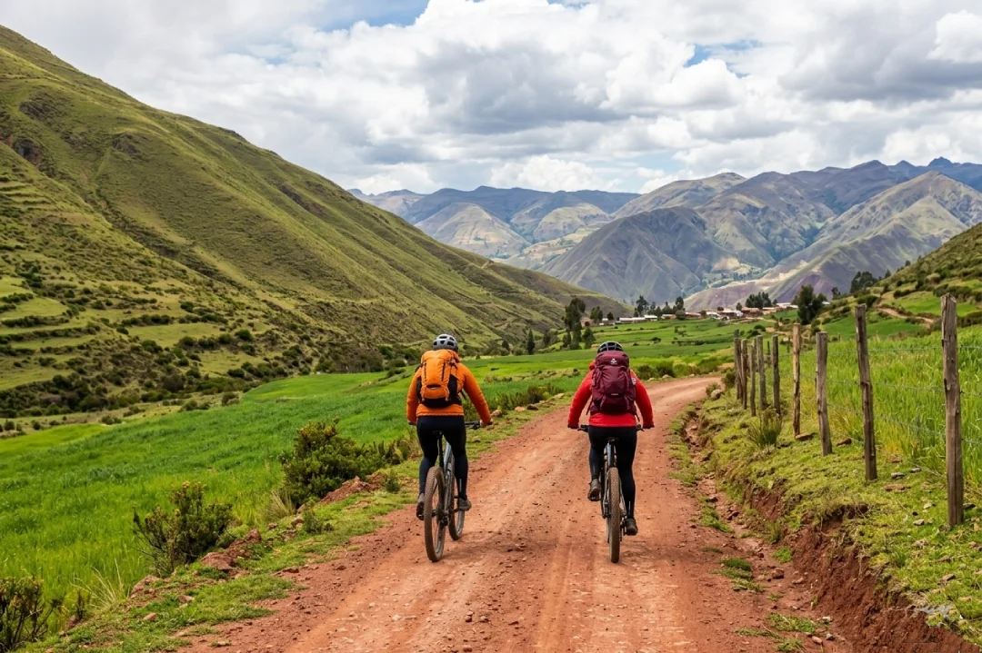 Dos ciclistas avanzando por un camino de tierra rojiza entre campos verdes en el Valle Sagrado, con montañas andinas y nubes dramáticas al frente.