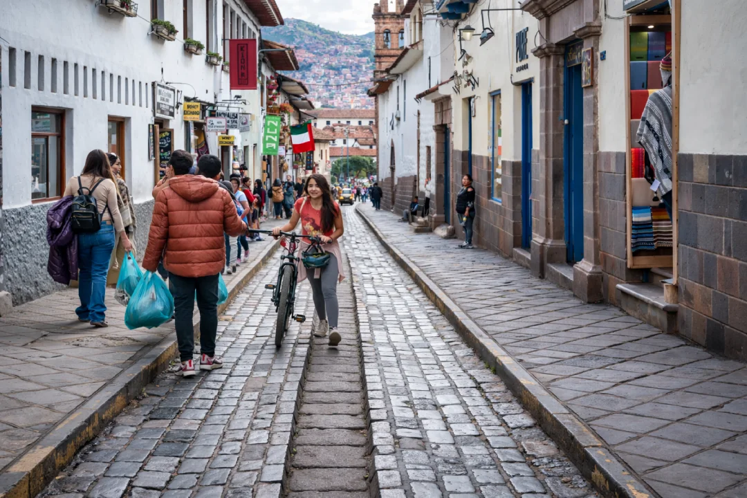 Mujer caminando con una bicicleta por una calle empedrada del centro histórico del Cusco, con peatones, casas coloniales y letreros al fondo.