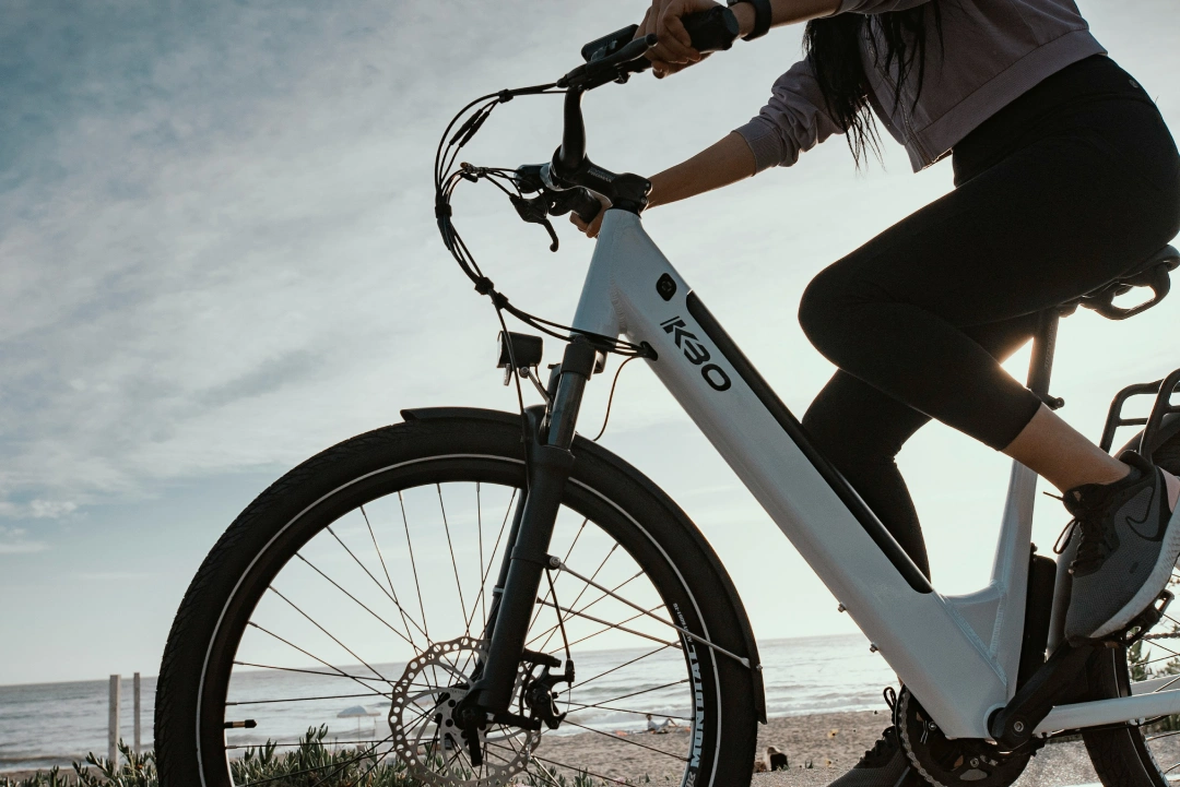 Low-angle photo of a rider on an electric bike near the beach at sunset, showing the frame, front wheel, and disc brake with ocean in the background.