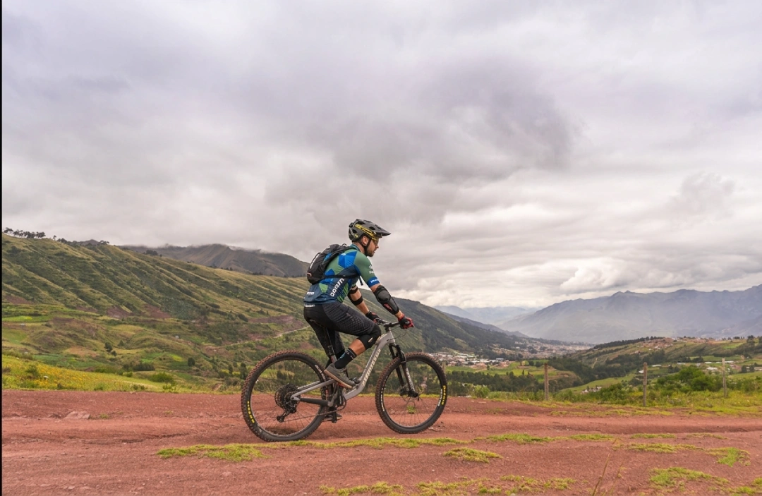 Mountain biker riding on a red dirt trail in Huayllarcocha near Cusco, with green hills and a wide valley view under cloudy skies. Huayllarcocha MTB route in Cusco