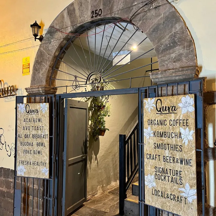 Entrada de Qura en Cusco con arco de piedra y letreros que anuncian café orgánico, kombucha, smoothies y desayuno todo el día.