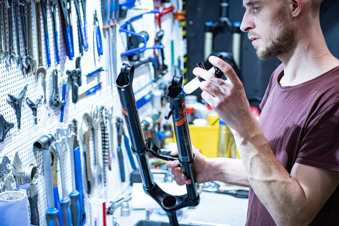 Bike mechanic in a workshop holding a suspension fork and a small bottle of fluid, with hand tools hanging on a wall in the background. How altitude affects MTB components
